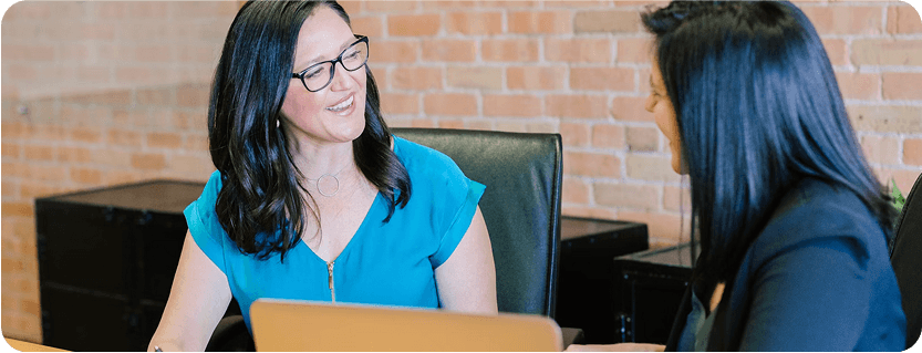 Two women chat smilingly at a work meeting in an office, with a laptop on the table.